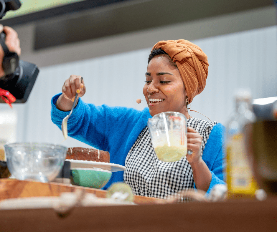Nadiya Hussain holding a jug of custard in one hand and spooning it onto a cake with the other, live onstage at BBC Good Food Show Summer