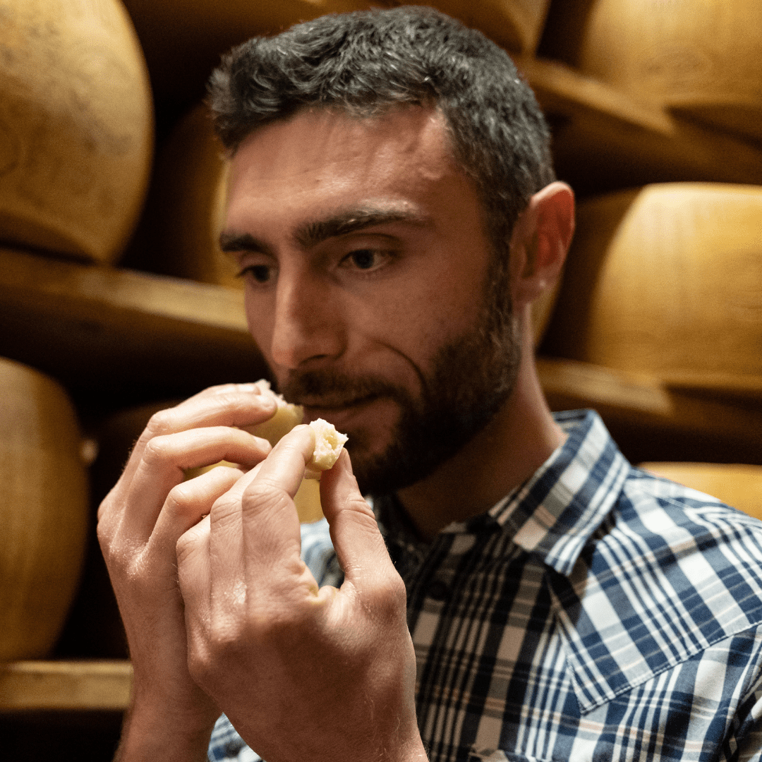 Alessandra Stocci smelling crumbs of cheese, with shelves of cheese wheels behind him